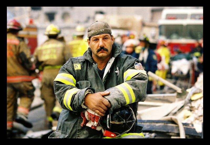 Color photograph of a firefighter sitting, resting with commotion of other firefighters and trucks behind him.